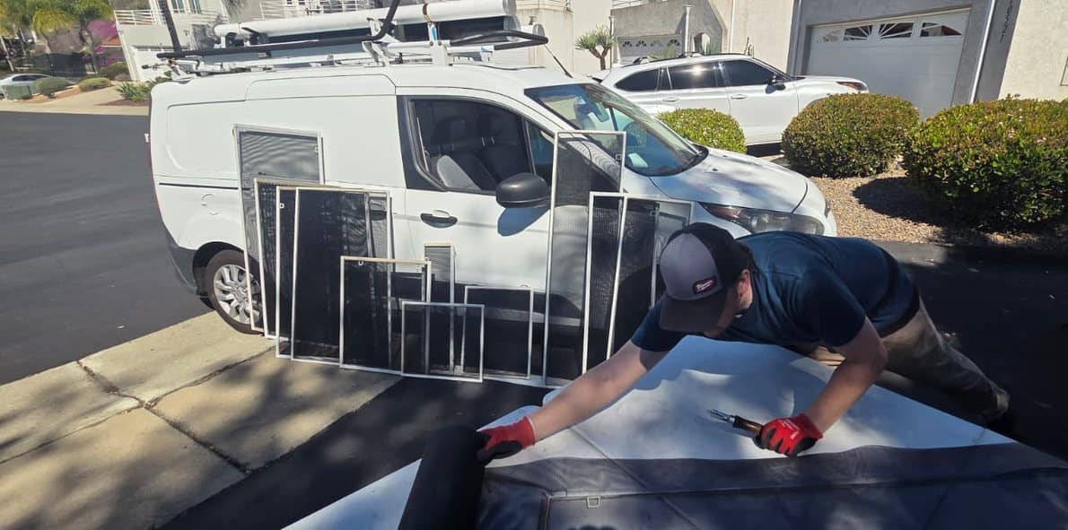 A technician setting new pet-resistant mesh into a window screen frame on a worktable in a driveway, with the service van parked behind